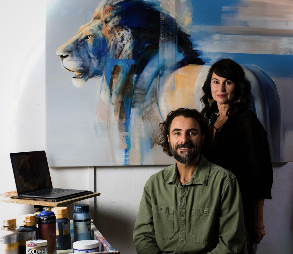 Das Künstlerpaar Frances Schandera-Duarte und Everett Duarte in ihrem Atelier. Im Hintergrund ist ein großformatiges, abstrakt-realistisches Gemälde eines Löwenkopfes zu sehen. Everett sitzt vor einer Staffelei mit Malutensilien, Frances steht dahinter.